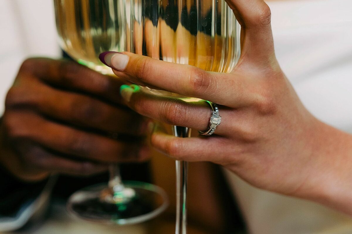 Image shows a newly wed couple toasting, with a focus on the trilogy set emerald engagement ring.
